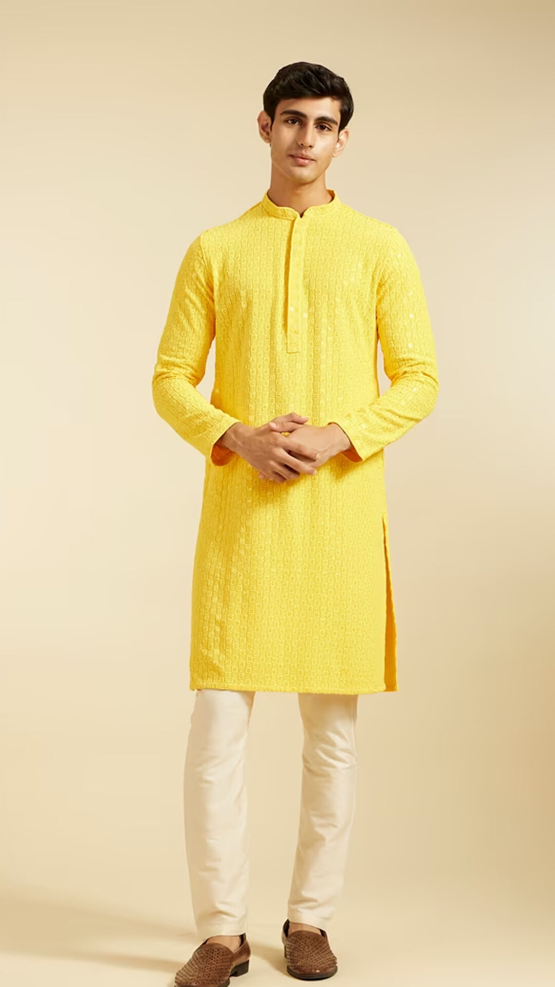 Man wearing a bright yellow textured kurta with cream churidar pants and brown traditional jutti, posing against a plain beige background